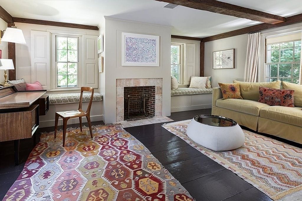 This cozy living room features a blend of rustic charm and eclectic decor, highlighted by exposed wooden ceiling beams and dark hardwood floors. The space is anchored by a central brick fireplace, flanked by built-in window seats and vibrant, patterned area rugs that add warmth and character. A comfortable sofa and a unique, modern coffee table create an inviting seating area, while a desk setup in the corner suggests a versatile use of the room.