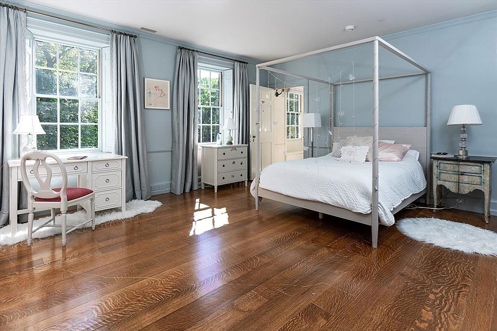 This elegant primary bedroom features a light blue color palette, hardwood flooring, and a prominent four-poster bed frame. The room is well-lit by natural light streaming through two large windows, which are framed by floor-to-ceiling curtains and accompanied by a dedicated desk area. The space exudes a serene and classic aesthetic, enhanced by soft white rugs and traditional furniture pieces.