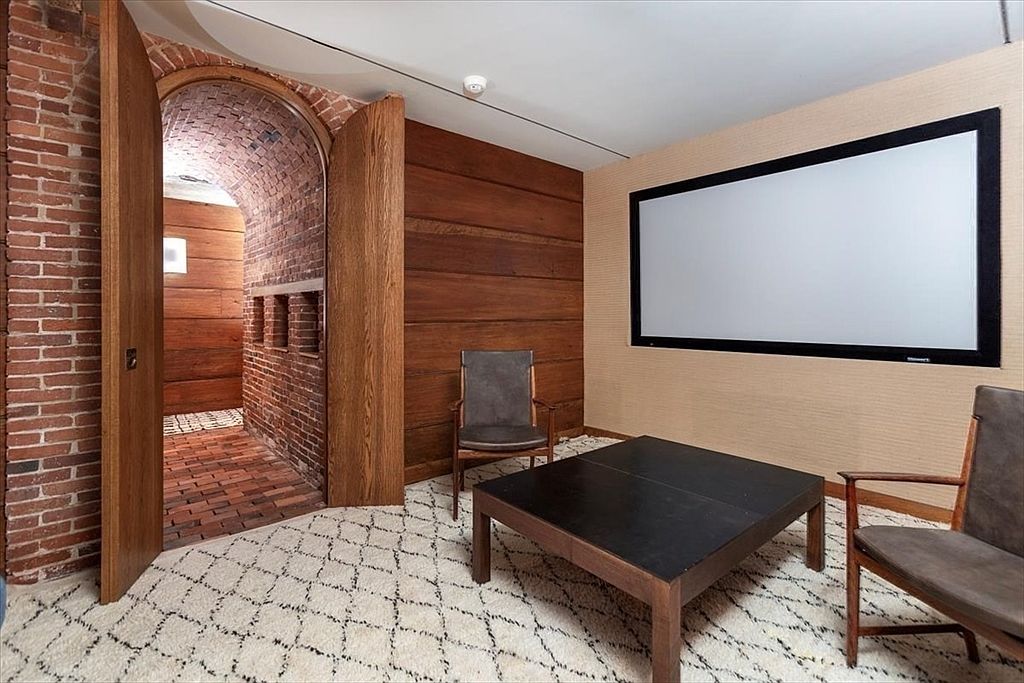 This unique basement media room features a striking arched brick entryway that leads into a rustic, tunnel-like corridor. The main room is furnished with mid-century modern leather chairs and a dark coffee table, set against a textured wall and a large projection screen. The combination of exposed brick, warm wood paneling, and a plush, patterned rug creates a cozy, cinematic atmosphere perfect for a private home theater.