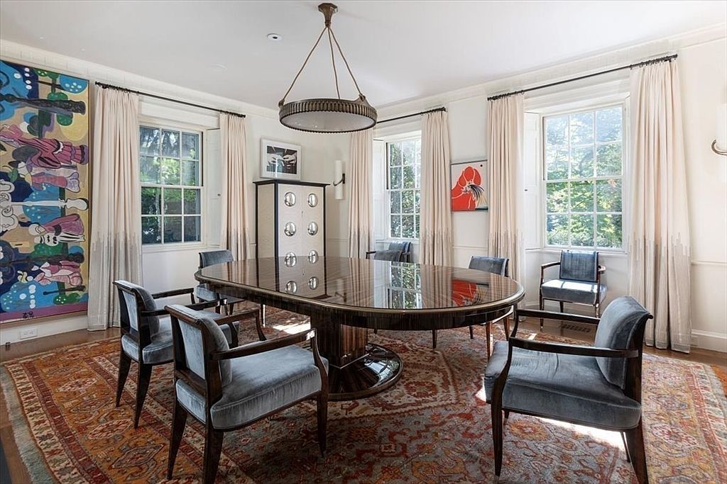 This elegant dining room features a large, polished oval table as its centerpiece, surrounded by six plush, velvet-upholstered armchairs. The space is anchored by a vibrant, traditional-style area rug and illuminated by a sophisticated, circular chandelier hanging from the ceiling. Large windows draped in floor-to-ceiling curtains allow natural light to fill the room, highlighting the eclectic artwork and the sleek, modern cabinet against the wall.