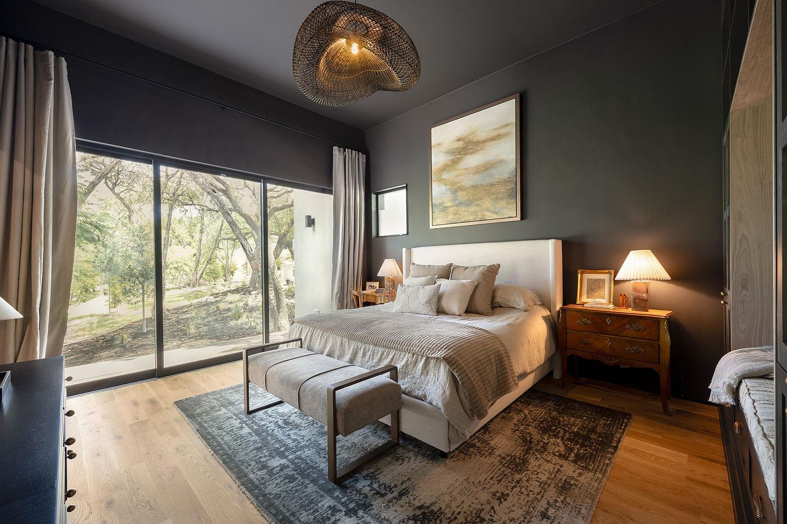 This is a primary bedroom featuring a large bed with neutral-toned linens and pillows, a bench at the foot of the bed, and dark gray walls. A large window provides natural light and a view of the outdoors, while a unique woven pendant light hangs from the ceiling. The room has a sophisticated and calming atmosphere.