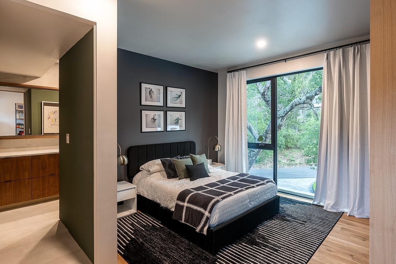 This is a primary bedroom featuring a dark accent wall adorned with four framed pictures. A black upholstered bed is neatly made with neutral bedding and decorative pillows, flanked by matching nightstands and lamps. Large windows with white curtains offer natural light and a view of the outdoors, while a striped rug adds texture to the hardwood floor.