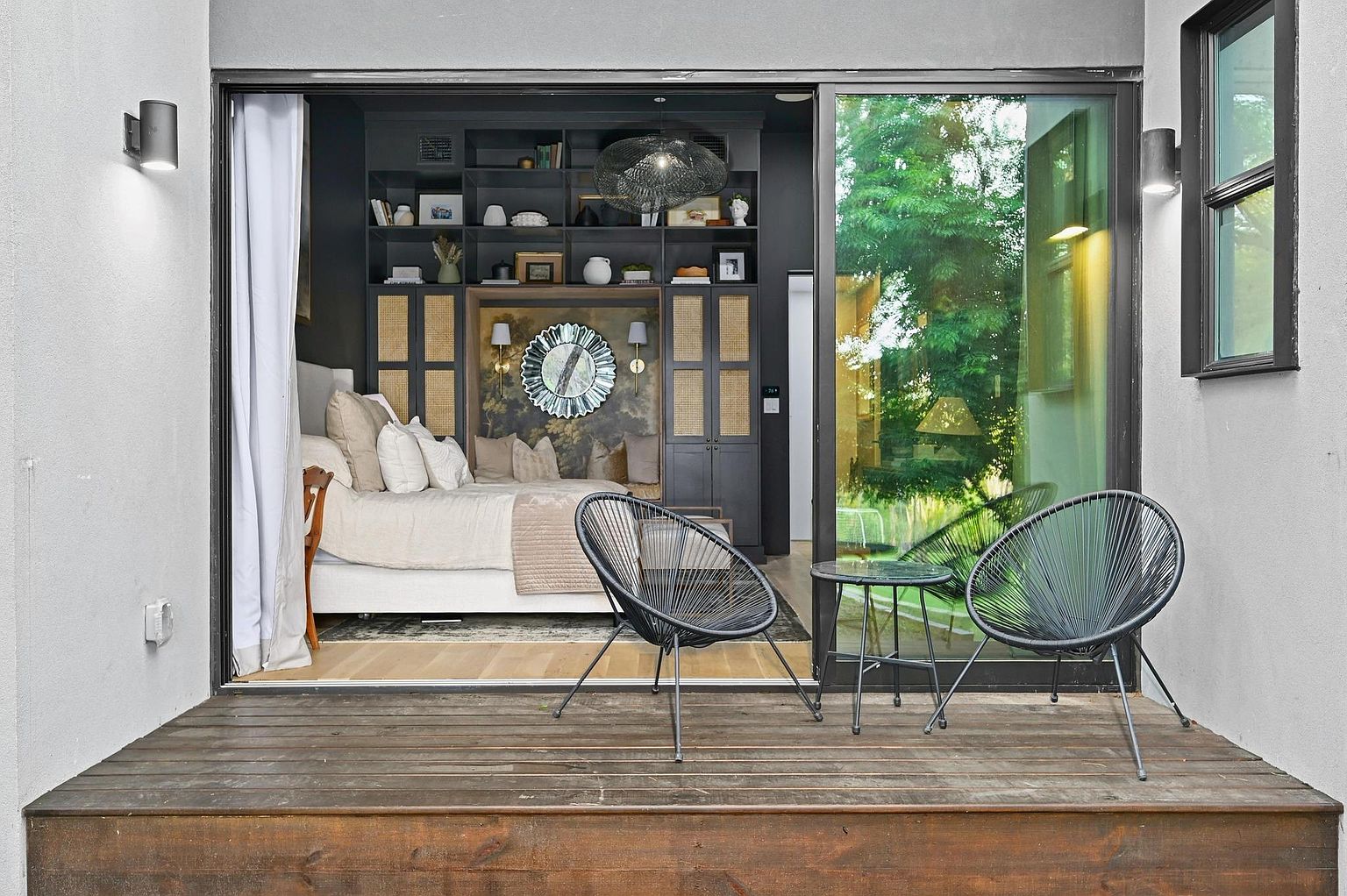 This image showcases a stylish patio or deck area seamlessly connected to an interior bedroom through large sliding glass doors. The outdoor space features a wooden deck, two modern black chairs, and a small glass-topped table, creating an inviting lounge area. The interior bedroom is visible, featuring a bed, built-in shelving, and decorative elements, blurring the lines between indoor and outdoor living.
