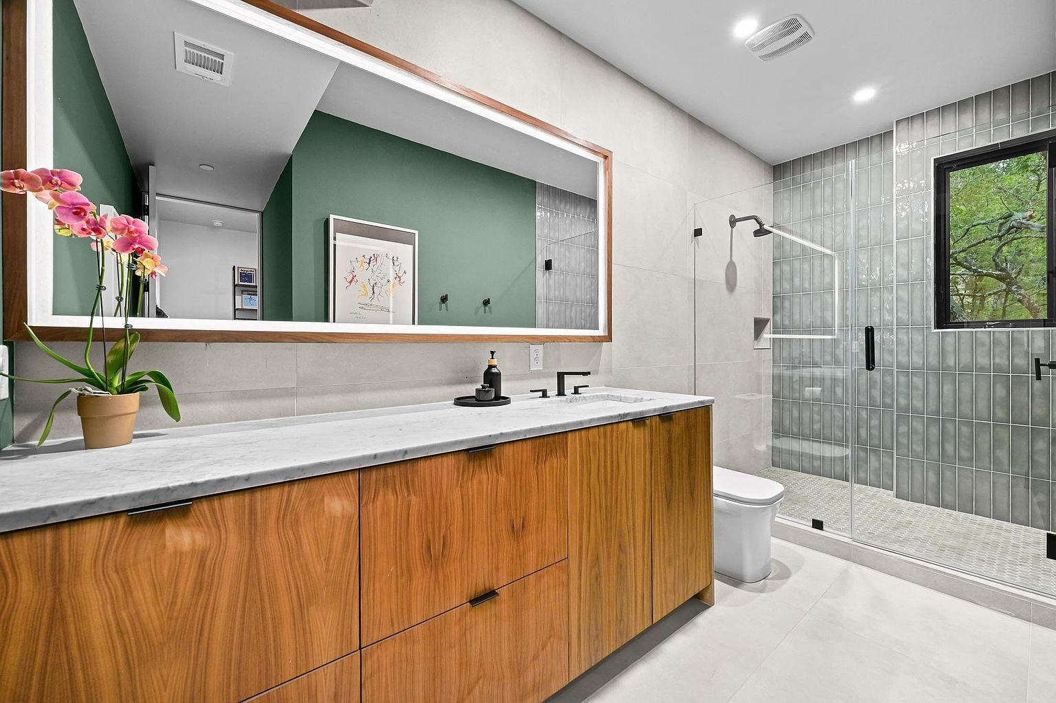 This is a modern primary bathroom featuring a wood vanity with a marble countertop, a large mirror with integrated lighting, and a glass-enclosed shower with gray vertical tiles. The bathroom has a clean and contemporary design with a neutral color palette, creating a spa-like atmosphere. A pink orchid adds a pop of color.