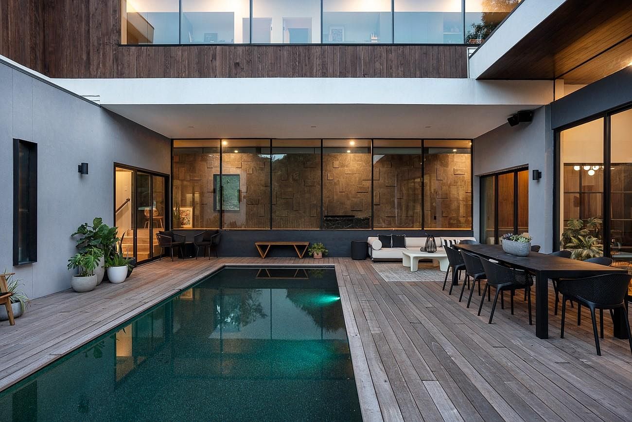 This image showcases a luxurious outdoor pool area, featuring a dark-bottomed pool surrounded by a wooden deck. The space includes an outdoor dining area with modern black chairs and a table, as well as a lounge area with a white sofa. Large windows provide a view into the home, creating a seamless indoor-outdoor living experience.