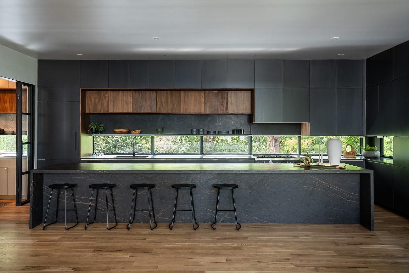 This is a modern kitchen featuring dark cabinetry, a large island with a unique stone countertop, and a long horizontal window providing natural light and a view of the outdoors. The kitchen island has seating for five, and the overall design is sleek and contemporary with a focus on clean lines and natural materials like wood and stone. The perspective is a wide shot, showcasing the entire kitchen area.