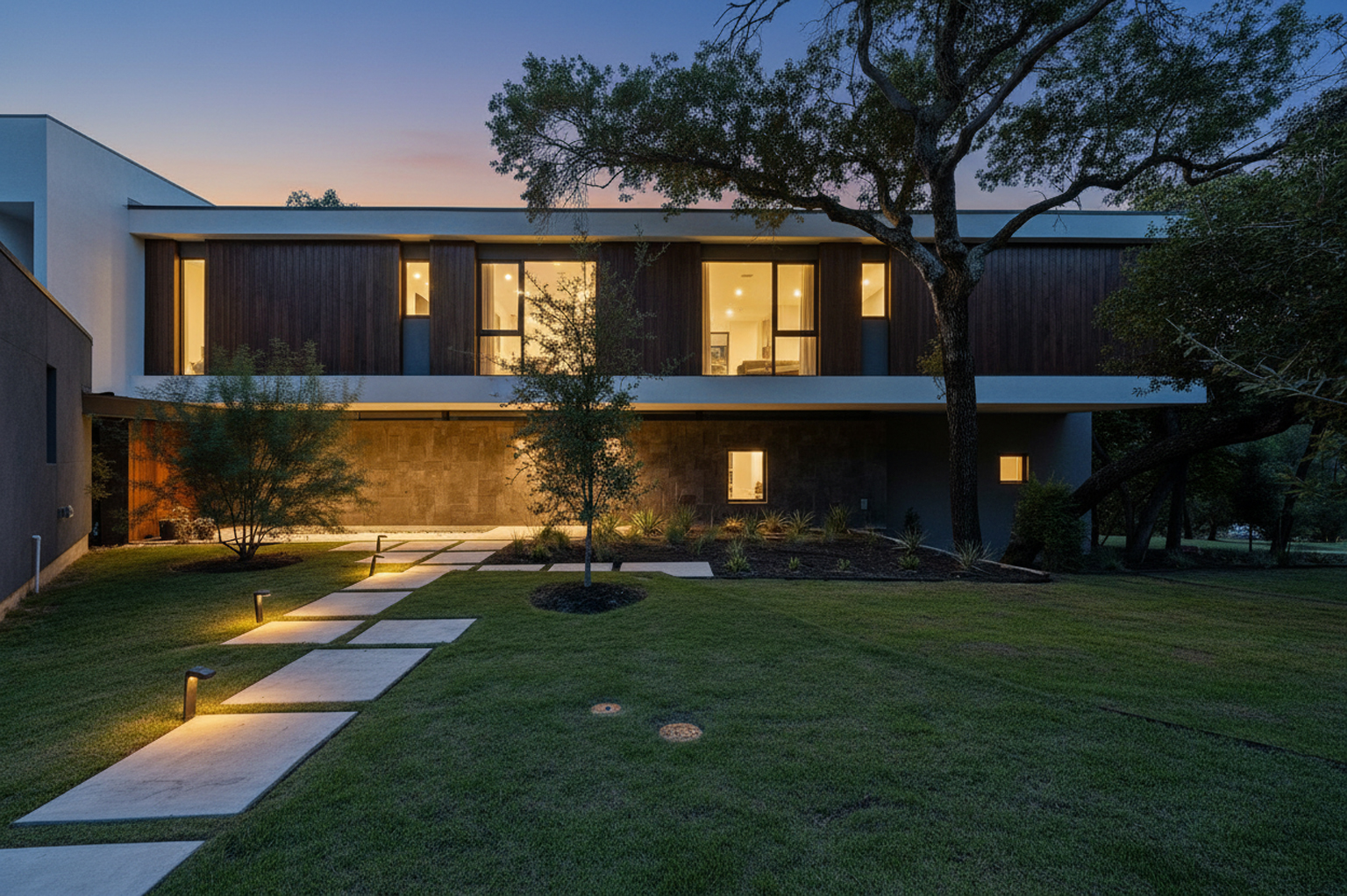 This image showcases the front exterior of a modern home at dusk, featuring a combination of wood and textured stone accents. A pathway of square concrete pavers, illuminated by landscape lighting, leads to the entrance. The well-maintained lawn and mature trees enhance the property's curb appeal.