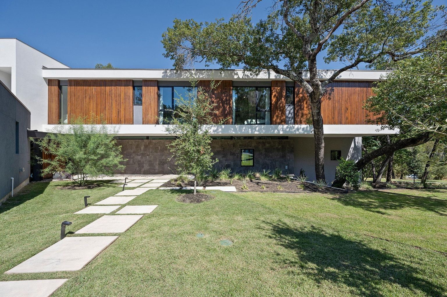 This is a front exterior view of a modern, two-story home featuring a combination of wood paneling, stone accents, and white stucco. Large windows provide ample natural light, and a concrete walkway leads to the entrance through a well-manicured lawn. Mature trees add to the property's curb appeal and provide shade.