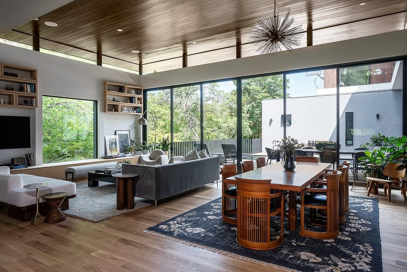 This is an interior shot of a modern living and dining area. The space features floor-to-ceiling windows offering a view of the outdoors, a gray sofa, a wooden dining table with unique chairs, and hardwood floors. The room is well-lit and has a contemporary design, creating a spacious and inviting atmosphere.