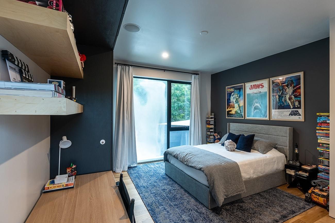 This is a well-decorated bedroom featuring a bed with a gray headboard, a blue patterned rug, and movie-themed posters on the wall. A desk with a lamp and shelves are on the left side of the image, while a stack of books and a toy robot are on the right. The room has a modern and playful aesthetic, suitable for a child or teenager.
