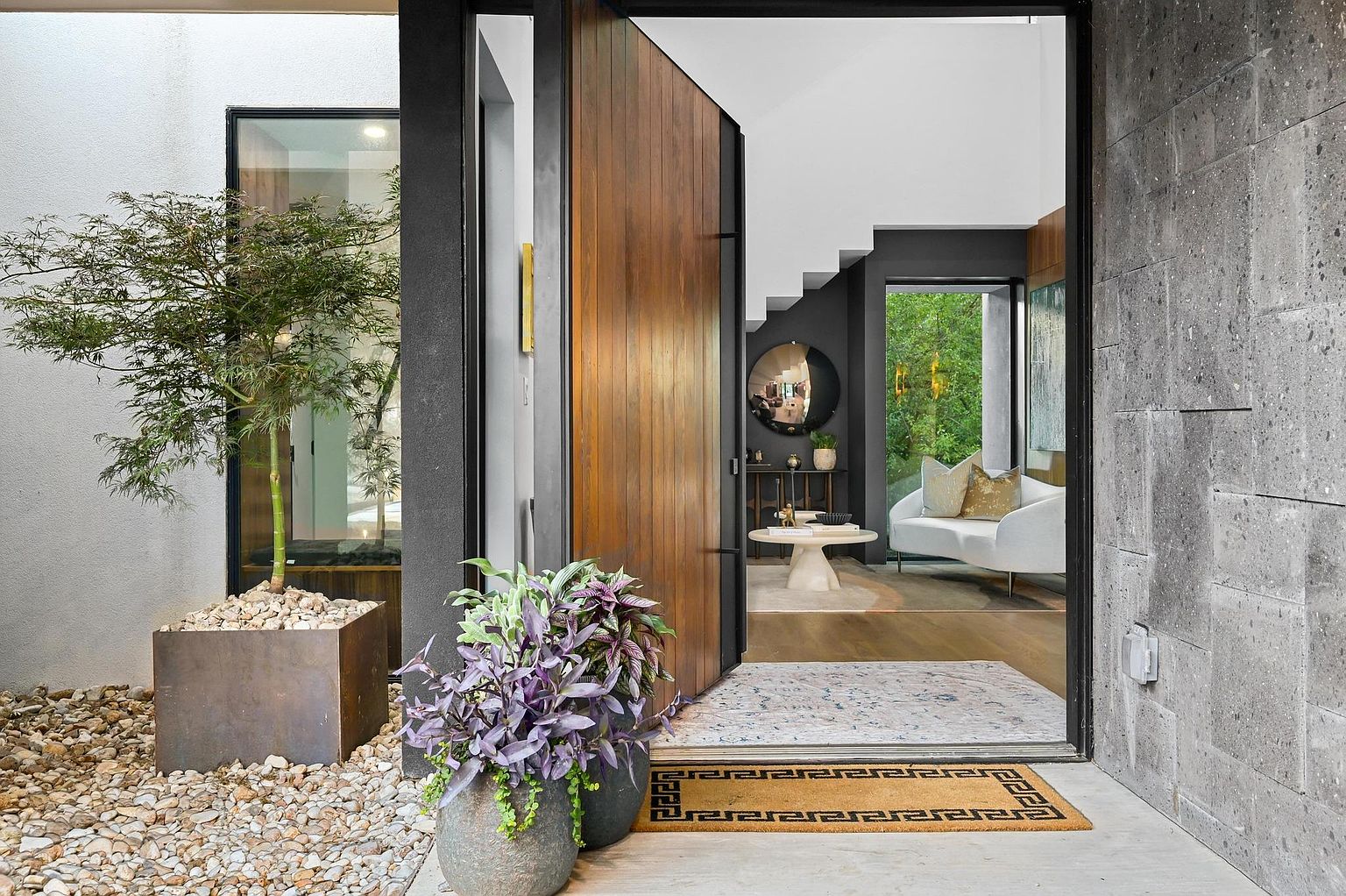 This image showcases a modern home's entryway, featuring a large wooden pivot door that is partially open, inviting entry. The exterior is accented with a small tree in a square planter, decorative rocks, and potted plants, creating a welcoming and stylish entrance. The interior visible through the doorway reveals a glimpse of a contemporary living space with a white sofa and modern decor.