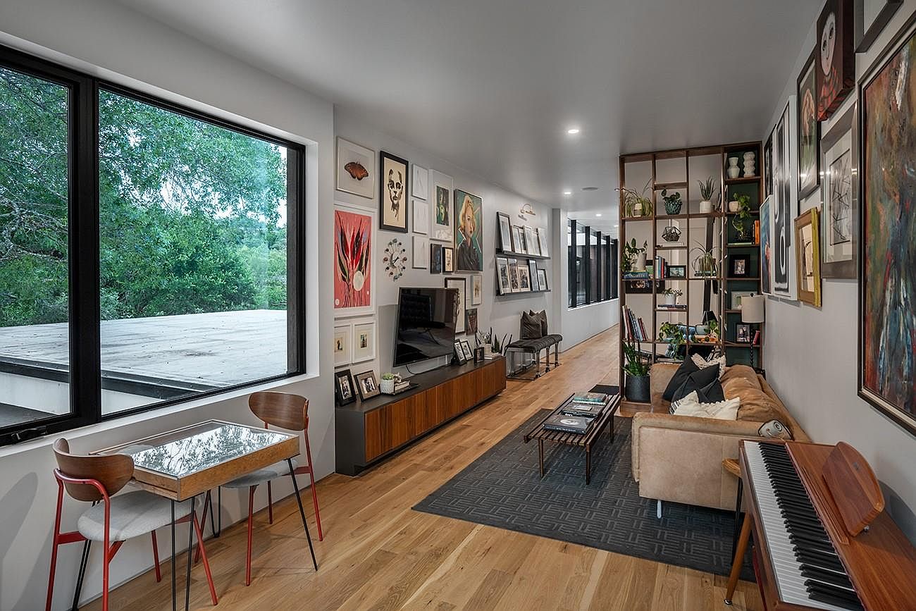 This is an interior shot of a modern living room featuring hardwood floors, a large window with a view of greenery, and a gallery wall with various framed artworks. The room is furnished with a sofa, a coffee table, a piano, and a media console, creating a stylish and inviting space. The perspective is from the side, showcasing the length of the room and the arrangement of furniture.