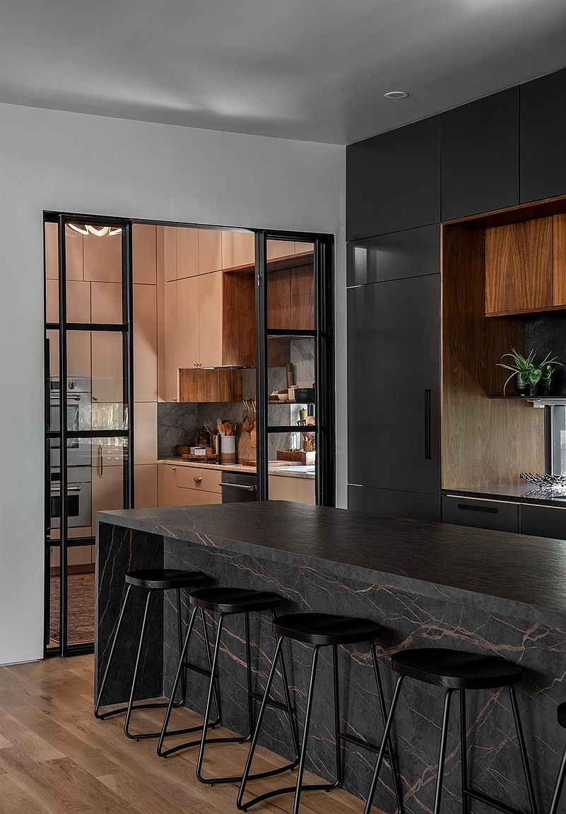 This is a modern kitchen featuring a dark marble island with black bar stools. The kitchen has a mix of dark and light wood cabinetry, with black framed glass doors leading to another kitchen area. The overall design is sleek and sophisticated, creating a luxurious and functional space.
