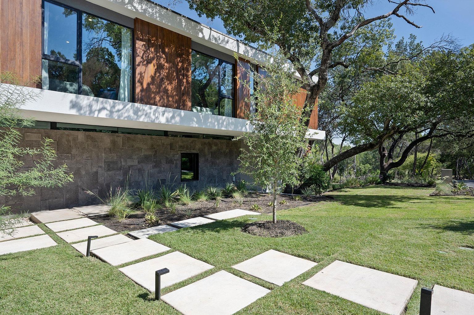 This image showcases the exterior of a modern home, focusing on the meticulously landscaped yard and garden. A pathway of square concrete pavers leads towards the house, flanked by lush green grass and carefully arranged plantings. The architectural style of the home is contemporary, featuring a combination of stone and wood cladding, with large windows reflecting the surrounding greenery.