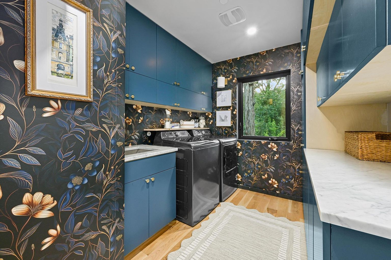 This is a well-appointed laundry room featuring dark blue cabinetry with gold hardware and a striking floral wallpaper. A modern, dark-colored washer and dryer set sits beneath a shelf, and a light-colored rug adds a touch of comfort to the hardwood floors. The room is illuminated by recessed lighting and natural light from a window, creating a stylish and functional space.