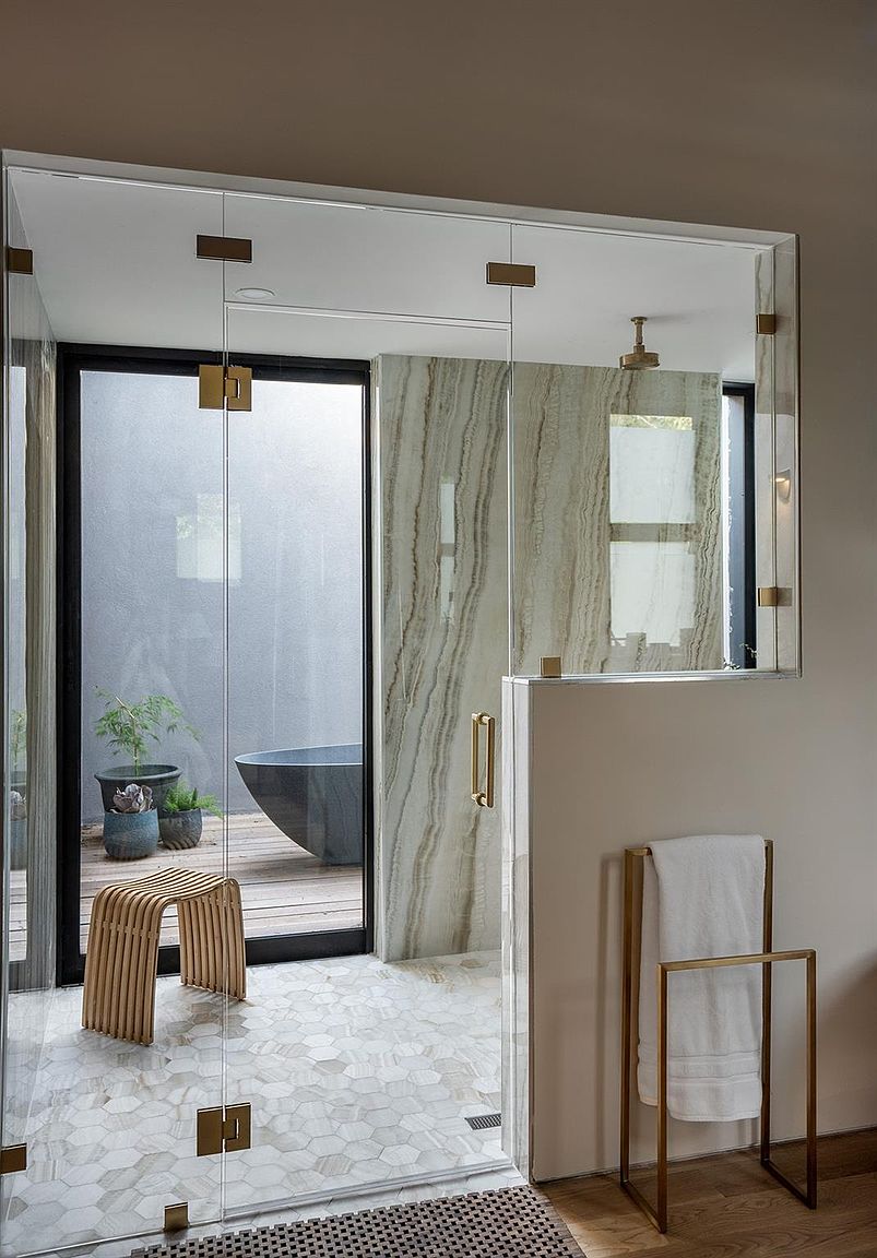 This is a luxurious primary bathroom featuring a glass-enclosed shower with marble-like walls and gold fixtures. The bathroom opens to an outdoor deck with a soaking tub and potted plants, creating a spa-like atmosphere. The interior flooring is a stylish hexagonal tile, and a wooden stool adds a touch of natural warmth.