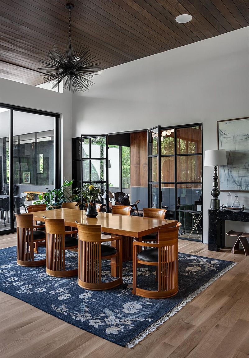 This is an interior shot of a dining room featuring a large wooden table surrounded by unique wooden chairs with black leather seats. A striking sputnik-style chandelier hangs above the table, and a blue floral rug anchors the space. Large windows and glass doors provide ample natural light, enhancing the room's modern and sophisticated aesthetic.