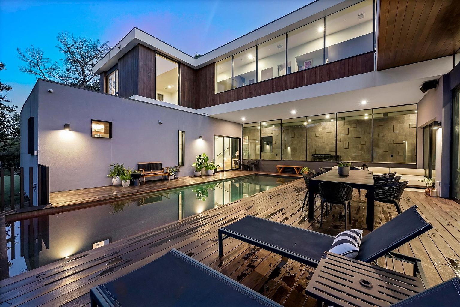 This image showcases a modern home's outdoor living space, featuring a sleek pool surrounded by a wooden deck. The deck is furnished with lounge chairs and a dining set, creating an inviting area for relaxation and entertainment. Large windows offer views into the home, blending indoor and outdoor living seamlessly.