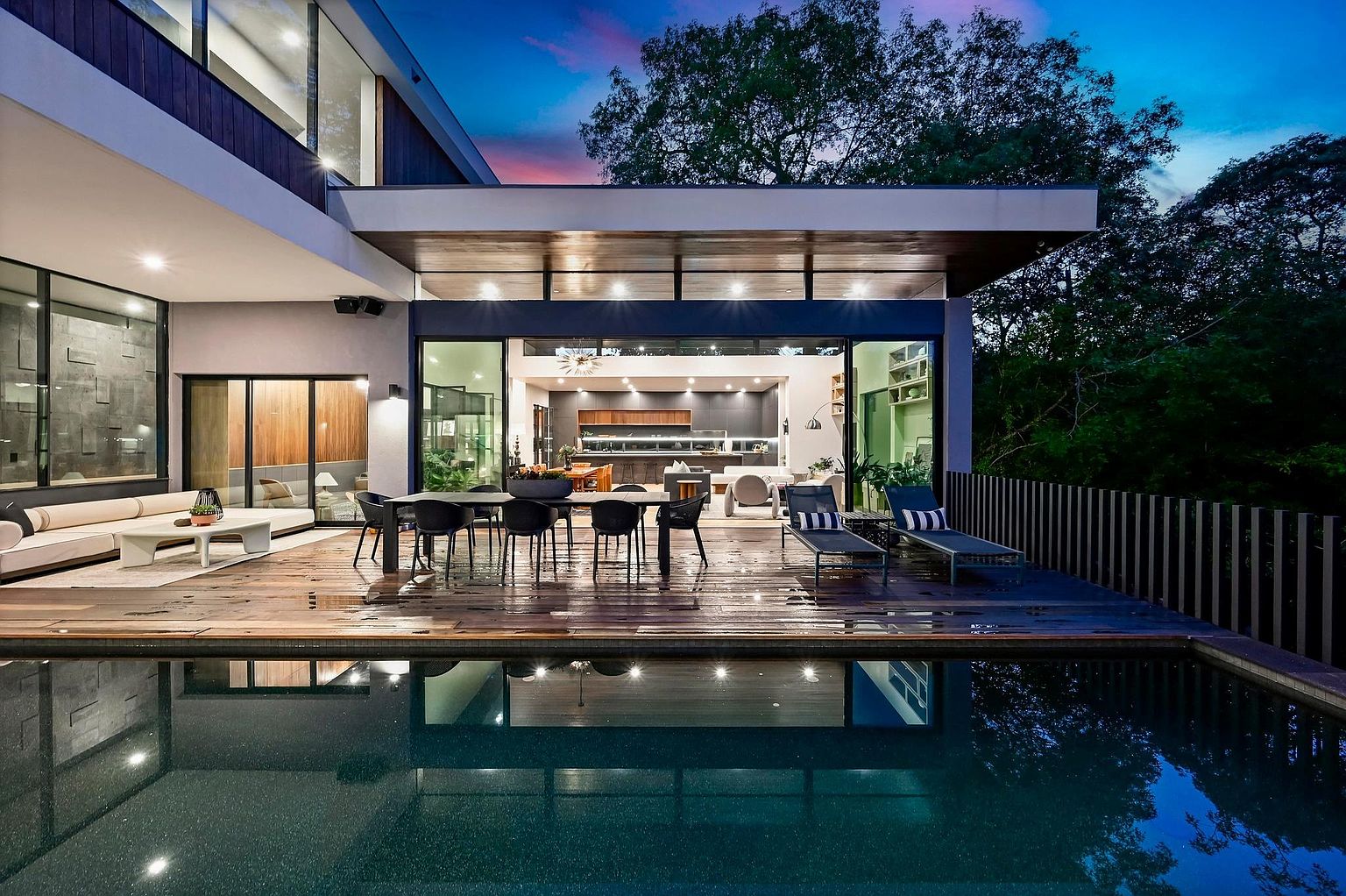 This is a stunning rear exterior view of a modern home featuring a wooden deck, outdoor dining area, and a luxurious pool. The open-concept design seamlessly connects the indoor kitchen and living spaces with the outdoor area, creating an ideal space for entertaining. The reflection of the house in the pool adds depth and visual appeal, showcasing the property's high-end design and inviting atmosphere.