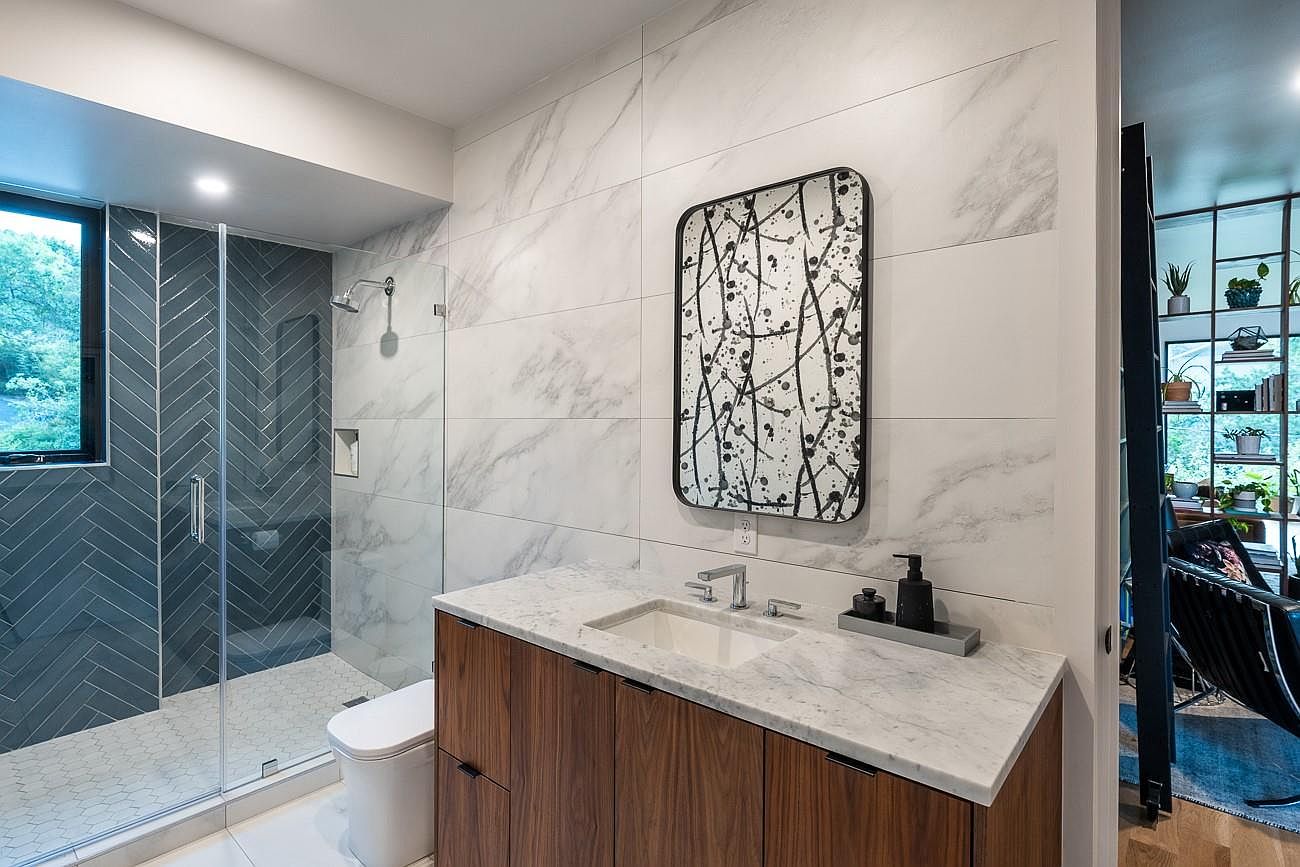 This is a modern bathroom featuring a glass-enclosed shower with blue herringbone tile, a white hexagon tile floor, and a floating wood vanity with a marble countertop. A contemporary mirror with an abstract design hangs above the sink, and the overall aesthetic is clean and stylish. The perspective is from the doorway, showcasing the entire bathroom.