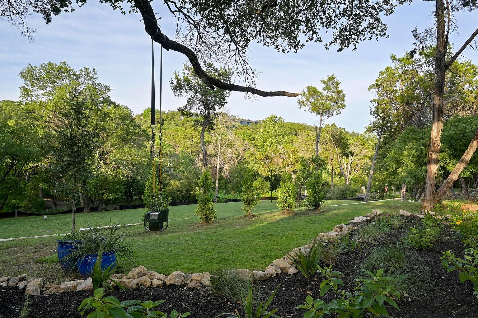 This image showcases a beautifully landscaped yard with lush green grass, mature trees, and carefully arranged rock borders. A unique tree swing hangs from a sturdy branch, adding a playful touch to the serene outdoor space. The scene is framed by a variety of plants and shrubs, creating a tranquil and inviting atmosphere.