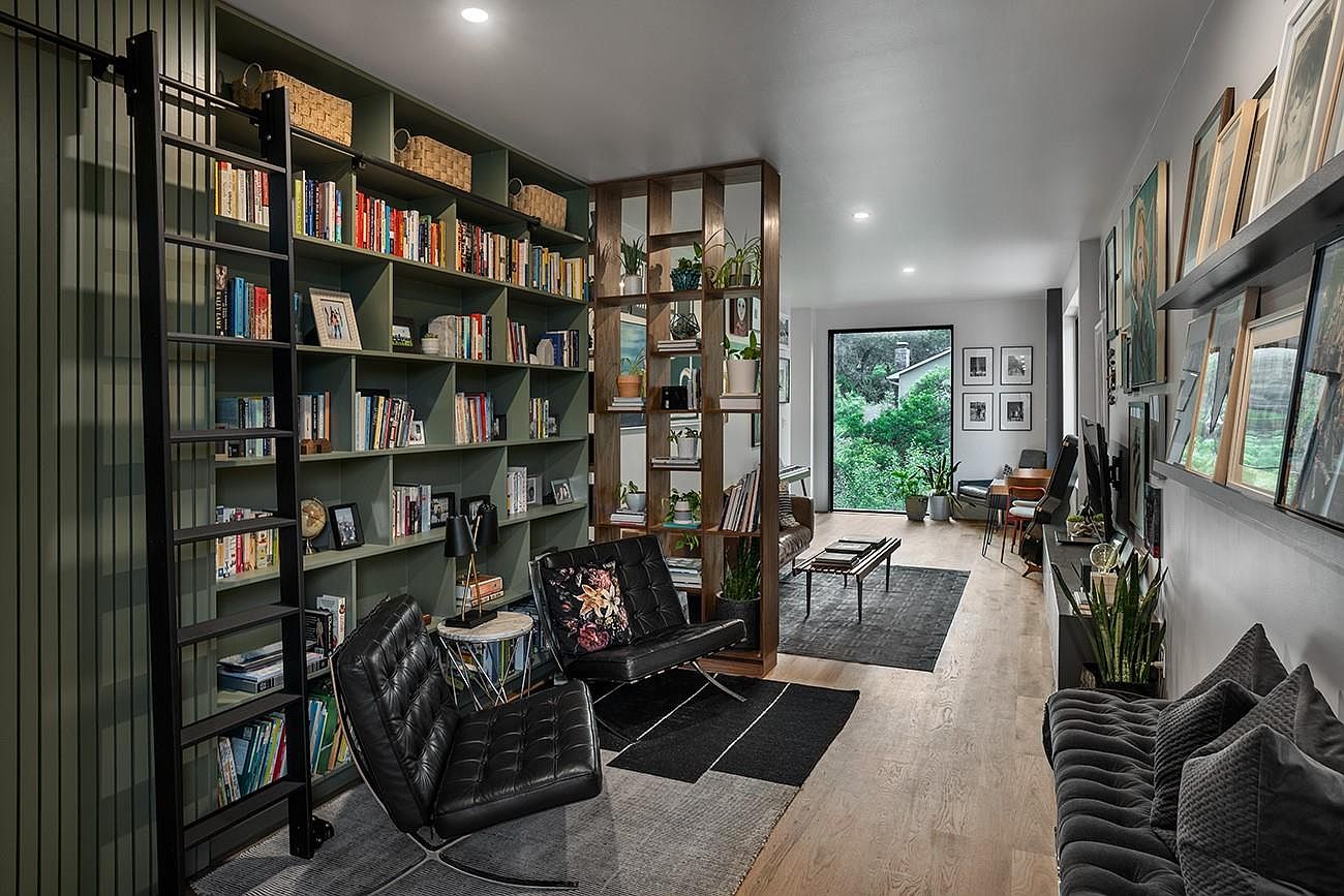 This is an interior shot of a stylish living room featuring a large bookshelf with a rolling ladder, two black leather chairs, and a wooden room divider. The room has hardwood floors and a gallery wall with framed pictures. A large window provides natural light and a view of the outdoors, creating a cozy and inviting atmosphere.
