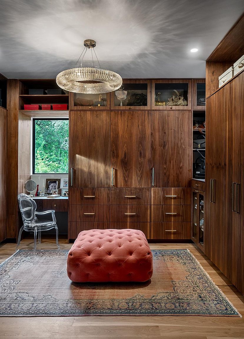 This is a well-organized walk-in closet featuring custom wood cabinetry with glass-fronted display sections and ample drawer space. A plush, tufted ottoman sits on a patterned rug, adding a touch of luxury. A vanity area with a clear acrylic chair is visible to the left, and a modern chandelier hangs from the ceiling, illuminating the space.