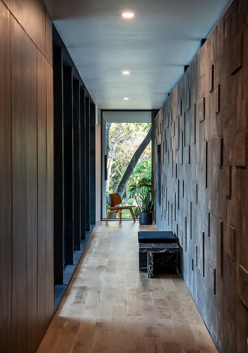 This interior shot showcases a modern hallway with unique architectural details. The hallway features a light wood floor, a textured wood panel wall, and a wall with vertical black slats. A chair and plant are visible at the end of the hallway, creating a sense of depth and inviting the viewer further into the home.