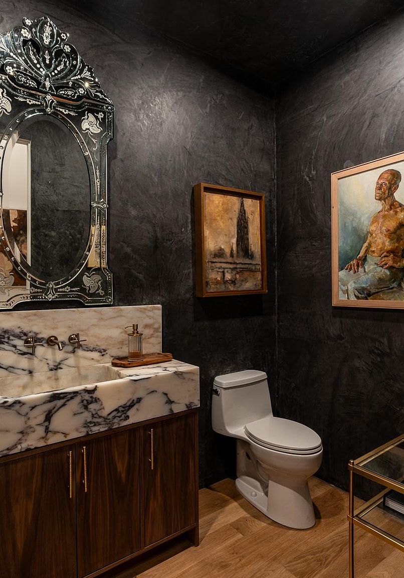 This is a stylish guest bathroom featuring dark textured walls, a marble countertop vanity with a dark wood cabinet, and an ornate mirror. The room is decorated with framed artwork, adding a touch of sophistication. The perspective is from the doorway, showcasing the vanity, toilet, and artwork.