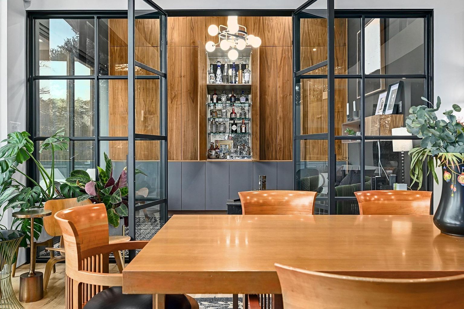 This interior shot showcases a dining room with a large wooden table and matching chairs, creating a warm and inviting atmosphere. Behind the dining area, there's a built-in bar with glass shelves and a modern chandelier, adding a touch of sophistication. The room is framed by black-framed glass doors, offering views of greenery and other interior spaces, enhancing the sense of openness and connection.