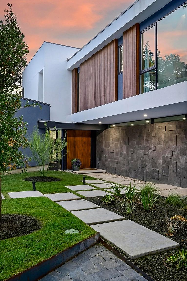 This image showcases the entryway of a modern home, featuring a striking combination of materials and textures. A wooden door is framed by a textured gray stone wall and sleek white architectural elements. The walkway consists of concrete pavers set amidst lush green grass and garden beds, creating an inviting approach to the residence.
