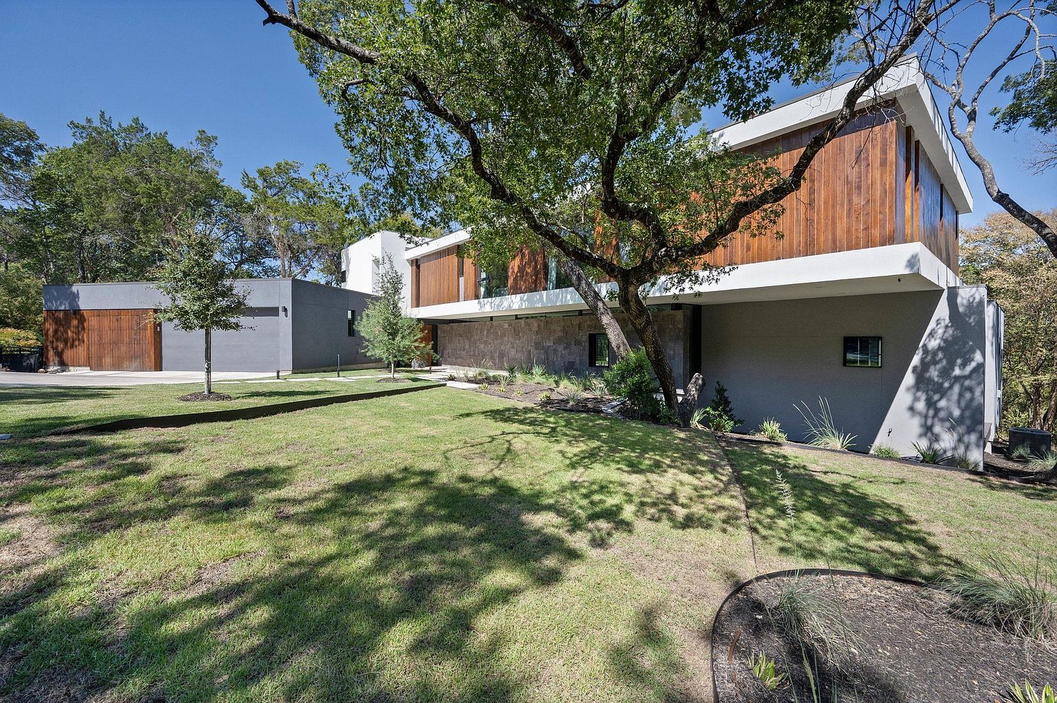 This is a front view of a modern, two-story home with a combination of gray stucco and wood paneling on the exterior. The house features clean lines, large windows, and a well-manicured lawn. A large tree in the front yard provides shade and adds to the property's curb appeal, while a detached garage complements the main structure.