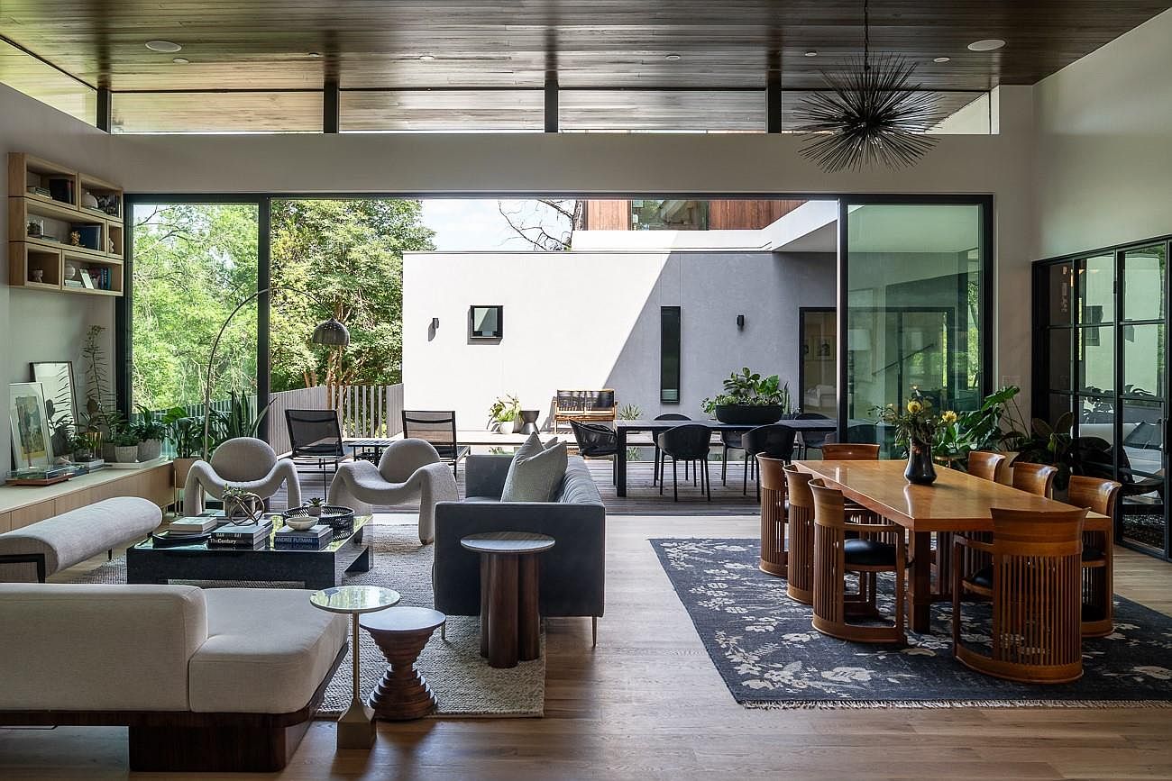 This is an interior shot of a modern living and dining area. The space features an open floor plan with large sliding glass doors that lead to an outdoor patio. The living area includes stylish seating arrangements, while the dining area showcases a wooden table with unique chairs, all set on hardwood floors with area rugs.