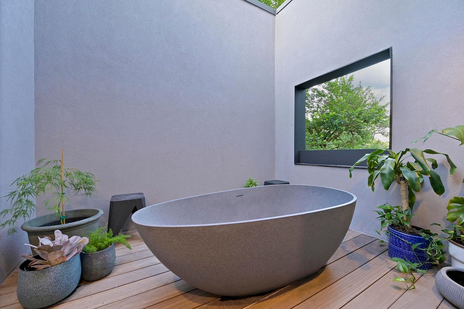 This image showcases a modern primary bathroom featuring a freestanding, oval-shaped bathtub as the focal point. The bathroom is designed with a minimalist aesthetic, incorporating a neutral color palette and natural light streaming through a strategically placed window. The wooden deck flooring and potted plants add a touch of warmth and nature to the serene space.