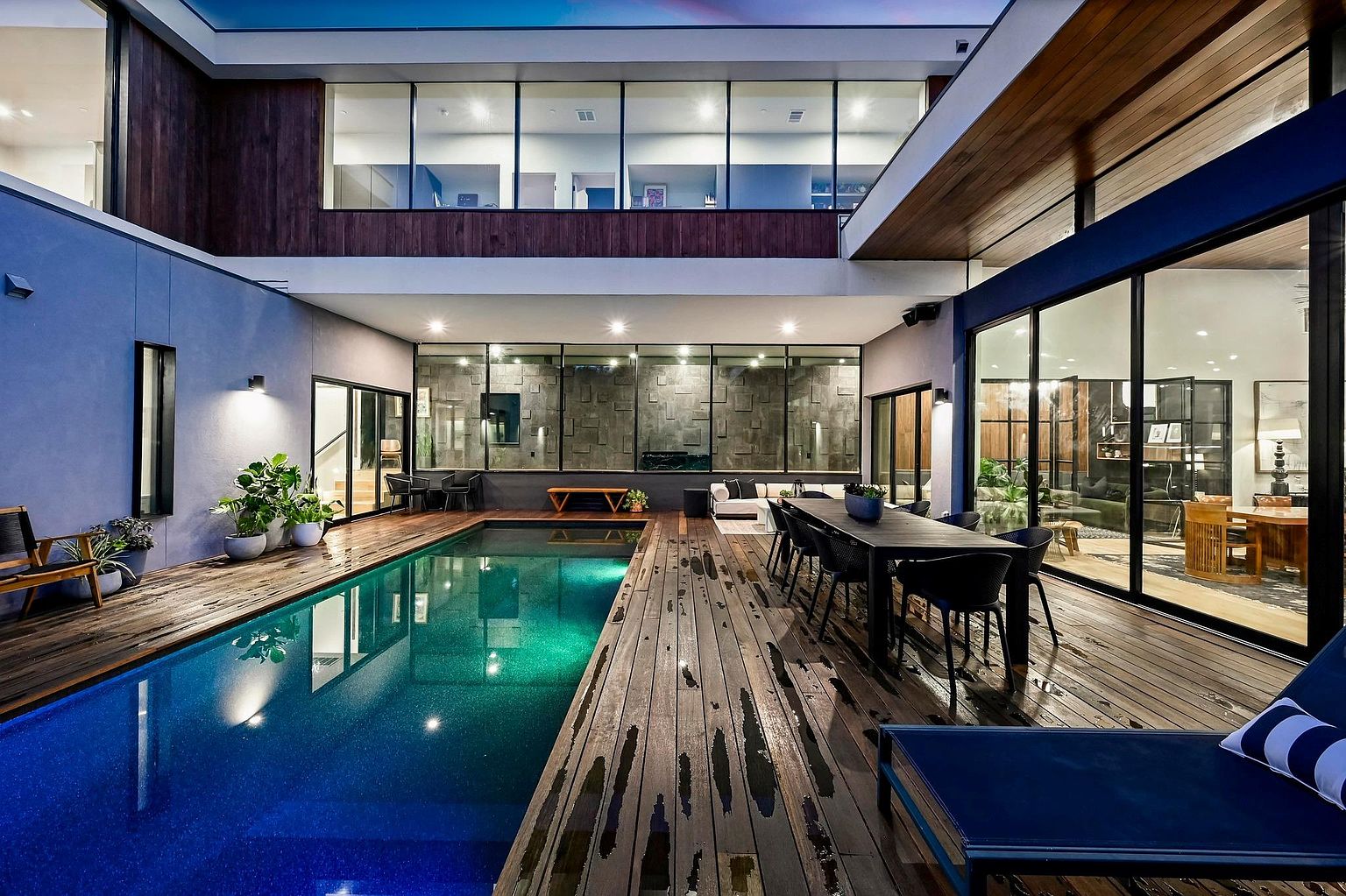 This image showcases a luxurious outdoor pool area, featuring a modern design with a wooden deck surrounding a vibrant blue pool. The space is enclosed by the home's architecture, with large windows offering views into the interior and creating a seamless indoor-outdoor living experience. A dining table and lounge chairs suggest the area is designed for both relaxation and entertainment.