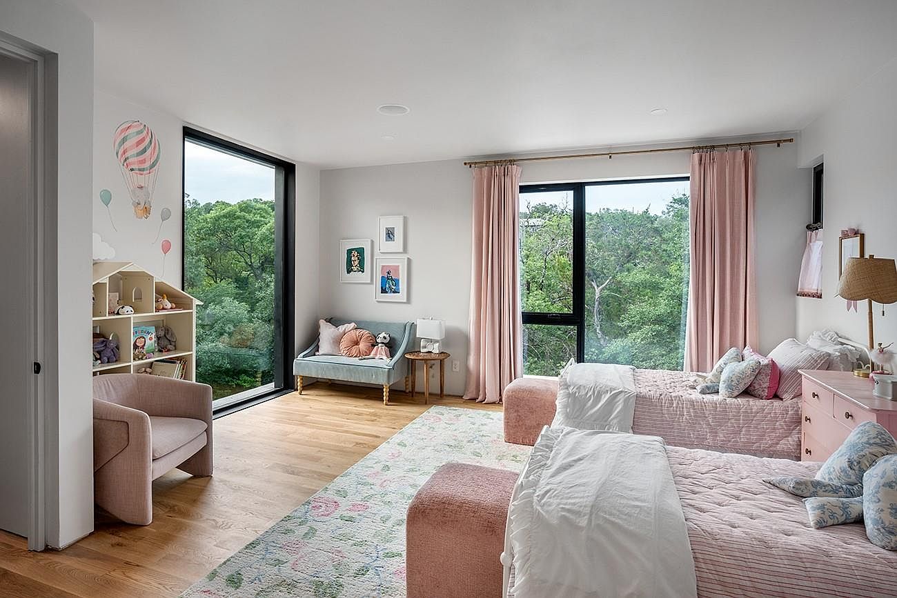 This is a charming children's bedroom featuring two twin beds with pink and white striped quilts and decorative pillows. The room is bright and airy, with large windows offering views of lush greenery. A small blue loveseat, a pink armchair, and whimsical wall decorations add to the room's playful and inviting atmosphere.
