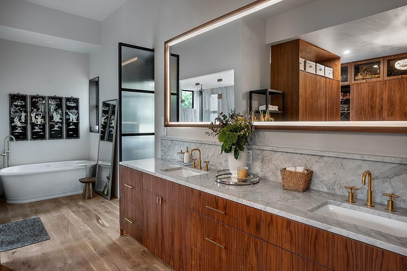 This is a luxurious primary bathroom featuring a double vanity with marble countertops and wood cabinetry. A large, illuminated mirror hangs above the vanity, reflecting the spacious layout. A freestanding bathtub is visible in the background, complemented by artwork and a separate glass-enclosed shower area, creating a spa-like atmosphere.
