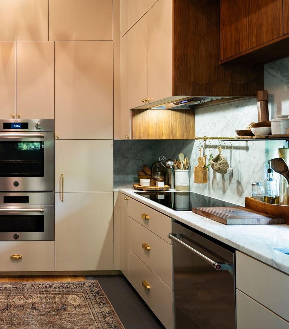 This is a well-appointed kitchen featuring a combination of light-colored cabinets with gold hardware and dark wood accents above the stove. Stainless steel appliances, including double ovens and a dishwasher, add a modern touch. The marble backsplash and countertops contribute to a luxurious feel, while the rug adds warmth and texture to the space.