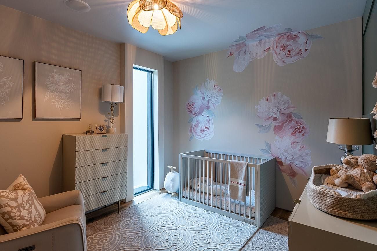 This is a charming nursery featuring a light gray crib with a personalized blanket, adorned with floral wall decals. The room is decorated in soft, neutral tones, complemented by a patterned rug and a unique flower-shaped ceiling light. A comfortable armchair and a dresser with a lamp add to the cozy and inviting atmosphere, perfect for a young child.