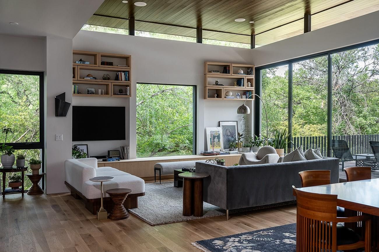 This is a bright and airy living room with floor-to-ceiling windows offering views of lush greenery. The room features a modern aesthetic with a gray sofa, a white chaise lounge, and wooden accents. Built-in shelving and a sleek TV add to the contemporary feel of the space.