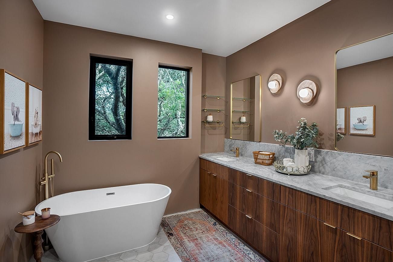 This is a primary bathroom featuring a freestanding white bathtub with gold fixtures, a double vanity with a marble countertop and wood cabinetry, and two windows providing natural light. The walls are painted in a warm, neutral tone, and the overall style is modern and luxurious. The perspective is from the doorway, showcasing the entire bathroom.