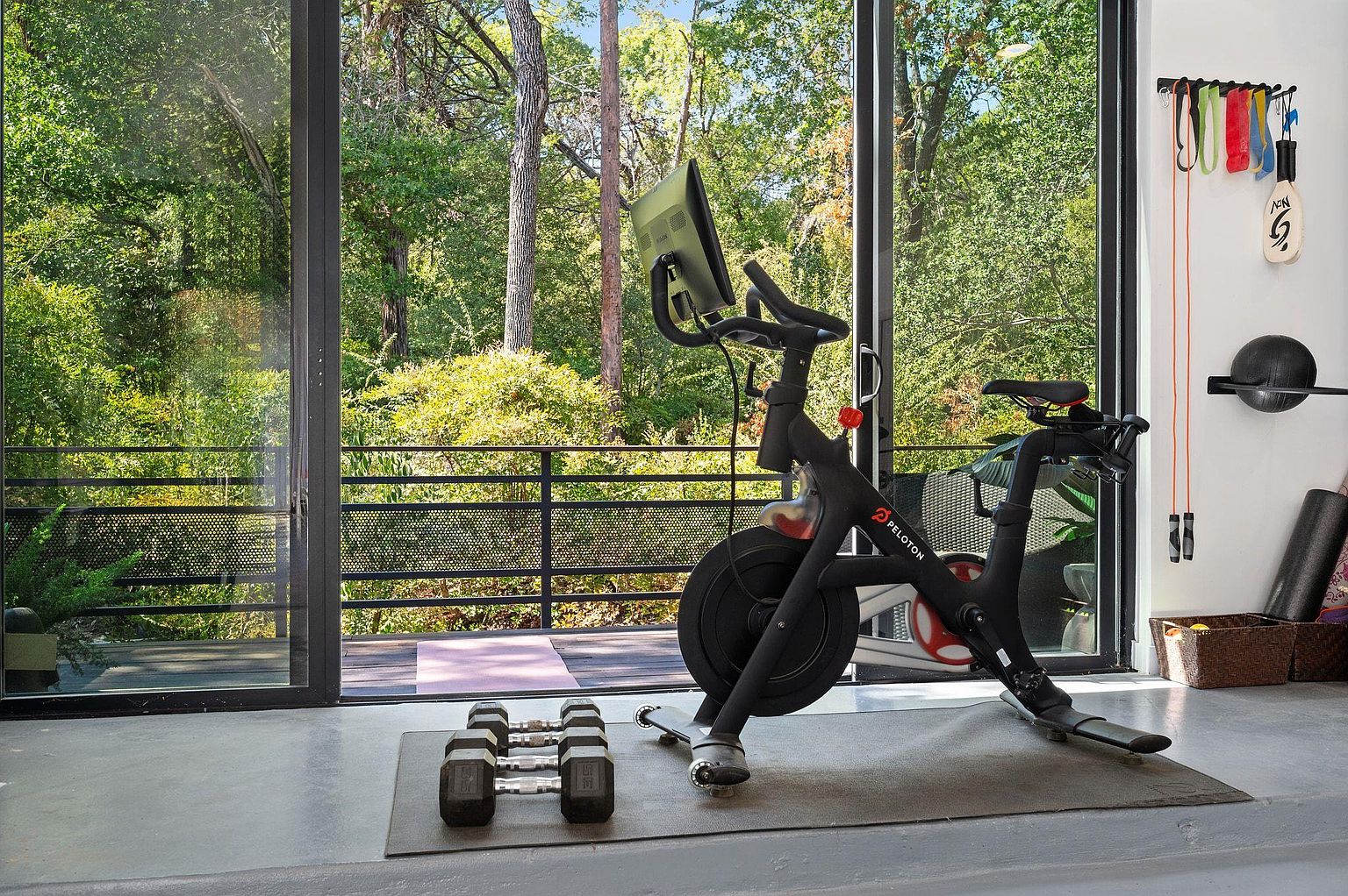 This interior shot showcases a well-equipped home gym with a Peloton bike as the focal point, positioned near large sliding glass doors that offer a view of a lush, green outdoor space. Dumbbells are neatly arranged on a mat, suggesting a dedicated workout area, while the natural light and serene backdrop create an inviting and motivating atmosphere for exercise.