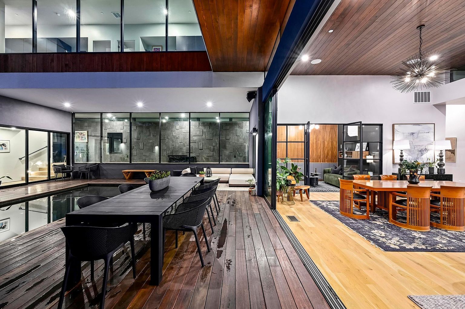 This image showcases a luxurious dining area that seamlessly blends indoor and outdoor spaces. The dining room features a large wooden table with unique chairs, complemented by a modern chandelier and artwork. Adjacent to the dining area is an outdoor space with a pool and a long dining table, creating an inviting atmosphere for entertaining.