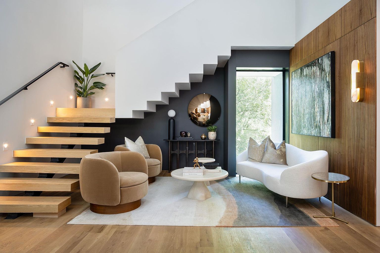 This is a stylish living room featuring a modern staircase with floating wooden steps and integrated lighting. The room is furnished with two round swivel chairs, a curved white sofa, and a round coffee table on a patterned rug. A large window provides natural light, and the overall aesthetic is contemporary and sophisticated.