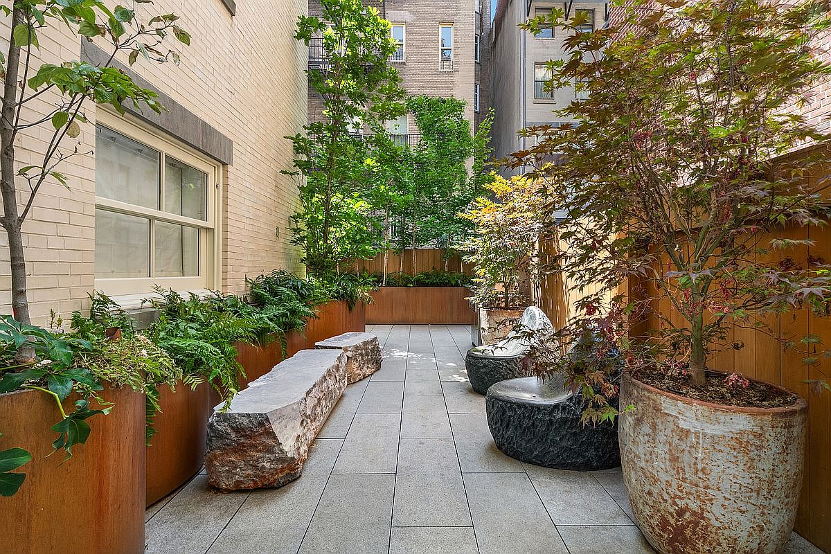 This sophisticated urban terrace features a clean, modern aesthetic with large stone pavers and custom metal planters filled with lush greenery. The space is furnished with unique, organic-shaped stone seating and a large decorative ceramic pot, creating a serene outdoor retreat nestled between brick buildings. The perspective is a low-angle, eye-level shot that emphasizes the depth and tranquil atmosphere of this private city garden.