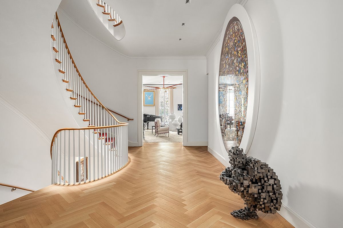 This elegant hallway features a curved staircase with a white balustrade and a light wood handrail, set against a backdrop of light-toned herringbone wood flooring. A striking, large oval mirror with a decorative, multi-colored mosaic frame hangs on the wall, complemented by a unique, geometric sculpture placed on the floor. The perspective looks through a doorway into a bright, adjacent room, creating a sense of depth and sophisticated architectural flow.