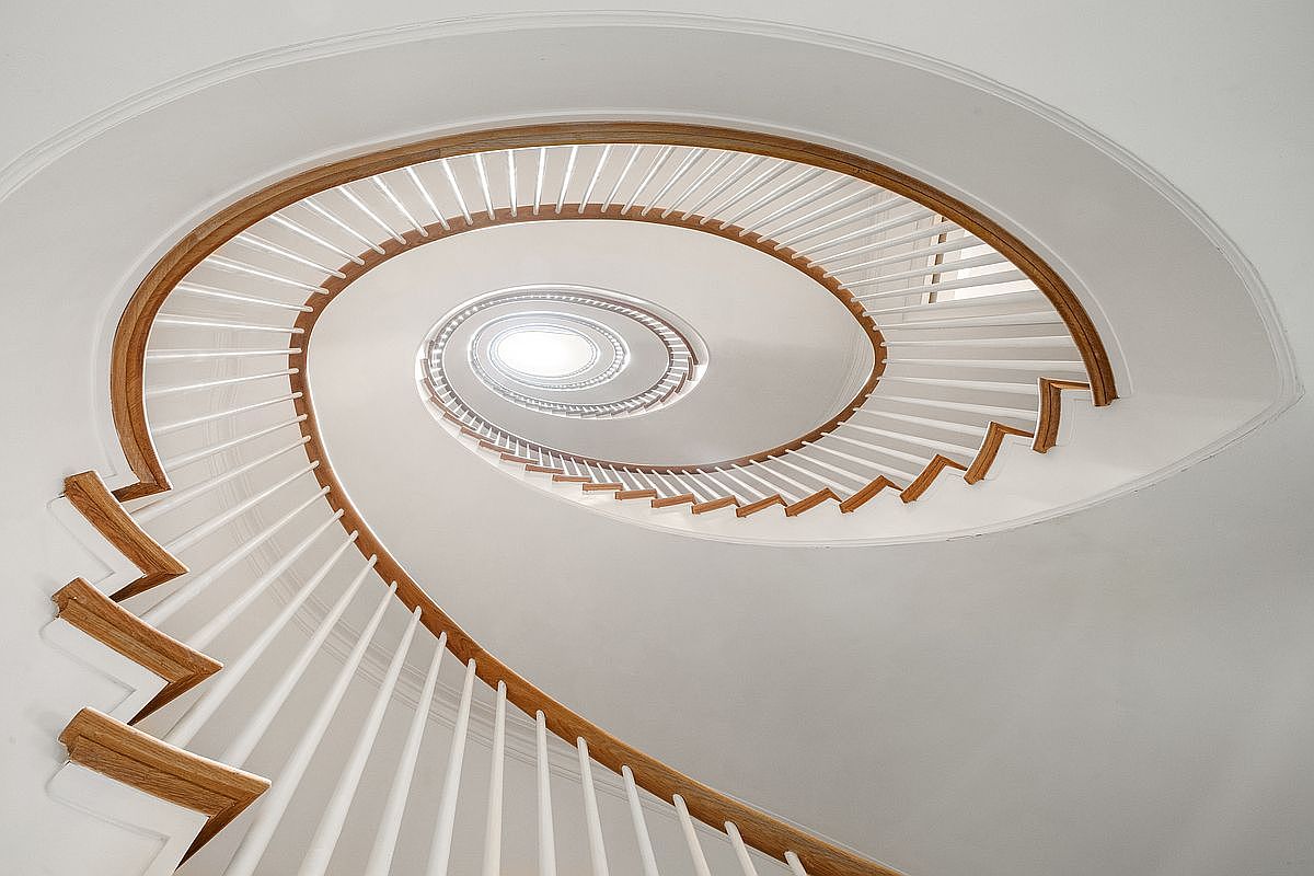 This image captures a dramatic, low-angle perspective looking up through the center of a grand spiral staircase. The design features elegant, light-toned wood handrails that contrast against crisp white balusters and walls, creating a clean, architectural aesthetic. The spiral leads the eye upward toward a bright, circular skylight at the apex, emphasizing the vertical scale and sophisticated geometry of the home.
