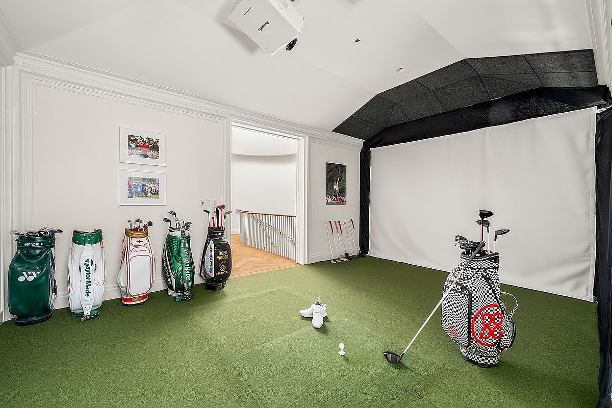 This dedicated indoor golf simulator room features a large projection screen, artificial turf flooring, and a ceiling-mounted projector. A collection of golf bags is neatly lined up against the wall, and a single golf club rests on the turf near a ball and a pair of shoes. The space is bright and clean, offering a professional-grade practice area for golf enthusiasts.