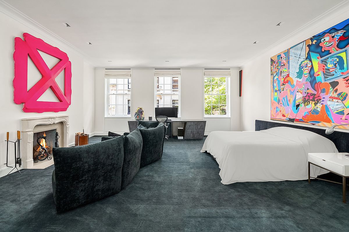 This expansive primary bedroom features a bold, contemporary aesthetic with a dark carpet and a large, vibrant abstract mural above the bed. A fireplace with a classic mantel serves as a focal point on the left, while a dedicated workspace with a desk and chair is positioned between two large windows. The room balances artistic flair with functional living spaces, creating a sophisticated and unique atmosphere.