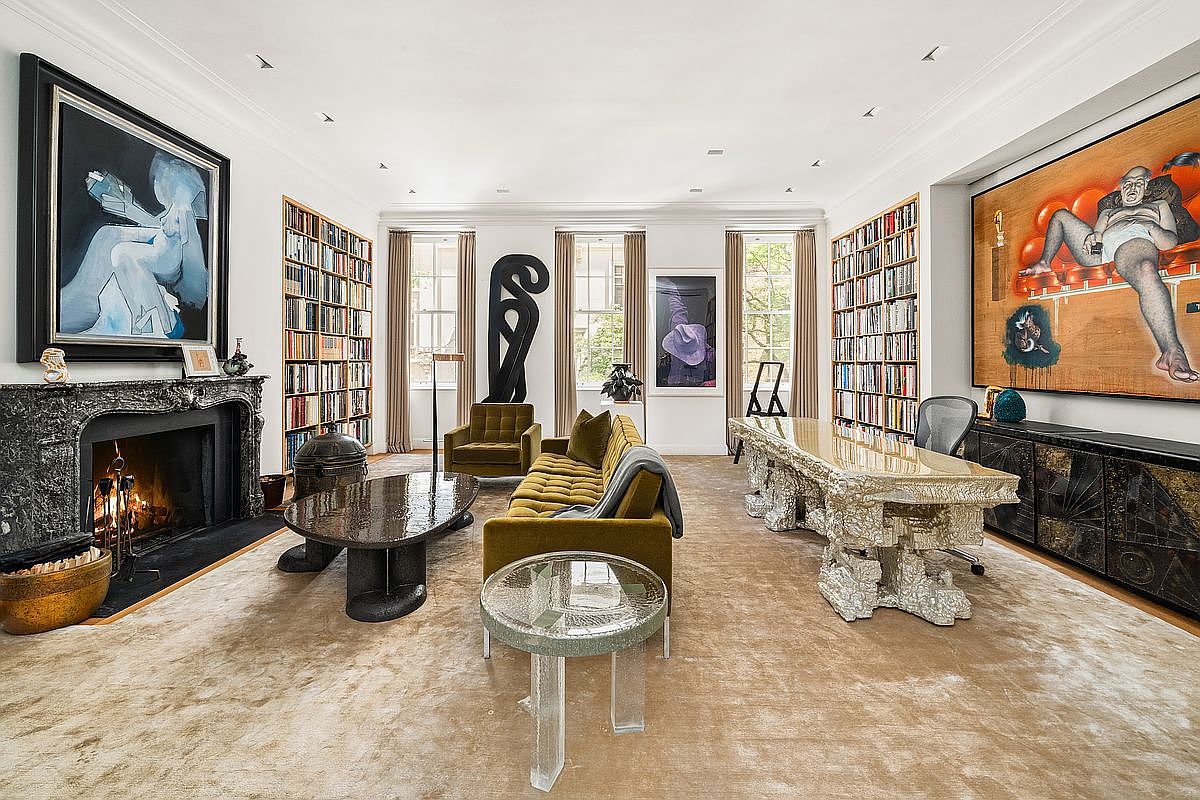 This sophisticated living room features a blend of eclectic art and high-end design, anchored by a dark stone fireplace surround and a plush, neutral-toned carpet. The space is furnished with a velvet sofa, a sculptural coffee table, and a unique, textured desk, all set against walls lined with extensive built-in bookshelves. Large windows with floor-length curtains allow natural light to fill the room, creating an atmosphere of refined luxury and artistic curation.